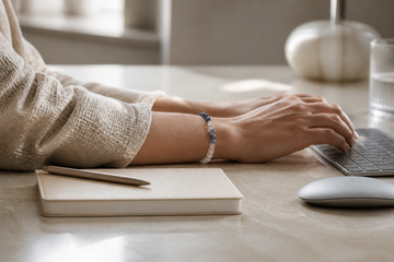 A restrained stone bracelet worn beside a keyboard, notebook, and neutral workwear